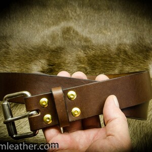 May include: A brown leather belt with a silver buckle and gold-colored rivets. The belt is held in a hand against a blurred background. The website address "tjpcustomleather.com" is visible at the bottom.
