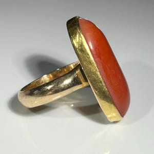 May include: Gold ring featuring a large, rectangular, orange-red stone. The stone is set in a gold bezel, and the band is also gold. The ring is displayed on a white surface.