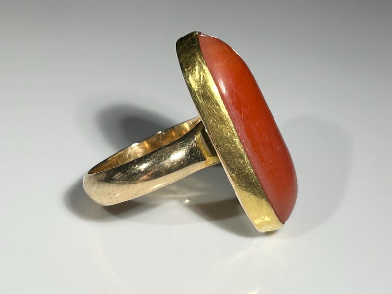 May include: Gold ring featuring a large, rectangular, orange-red stone. The stone is set in a gold bezel, and the band is also gold. The ring is displayed on a white surface.