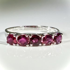 May include: A silver band ring featuring five round, faceted, deep pinkish-red gemstones set in a row. The metal band has a polished, reflective finish, and the stones are secured by small silver prongs, creating a continuous line of color.