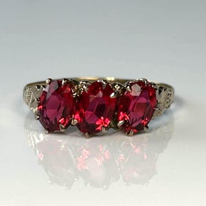 May include: A gold-coloured ring with three oval-cut, bright red gemstones. The ring's band has a decorative floral design on the sides. The gemstones are set closely together, creating a striking visual effect. The ring is displayed on a reflective surface.