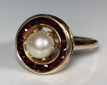 May include: A gold ring with a white pearl surrounded by a halo of red gemstones.