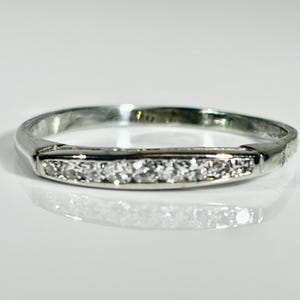 May include: A silver-toned metal band ring featuring a central horizontal bar set with a row of seven small, clear, round-cut stones. The ring has a polished, reflective finish and a slim, uniform band that tapers slightly toward the back.