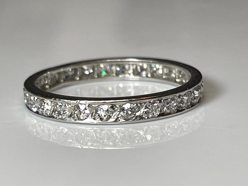 May include: A silver band ring featuring a continuous row of small, clear gemstones. The ring is set against a white background, reflecting light. The ring's design is simple and elegant, suitable for everyday wear.