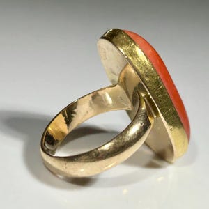 May include: A gold ring with a large, oval-shaped coral stone. The coral is a vibrant orange-red color, set in a gold bezel. The ring band is also gold, with a smooth, polished finish.