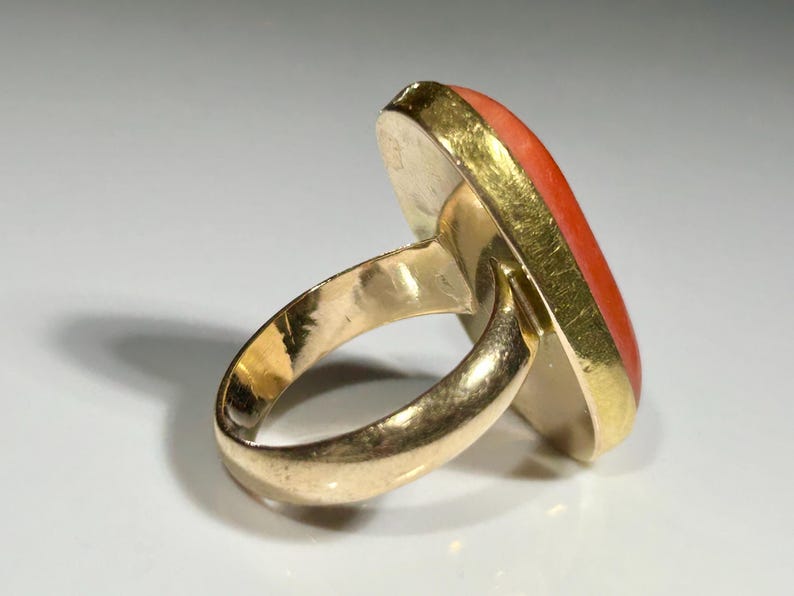May include: A gold ring with a large, oval-shaped coral stone. The coral is a vibrant orange-red color, set in a gold bezel. The ring band is also gold, with a smooth, polished finish.
