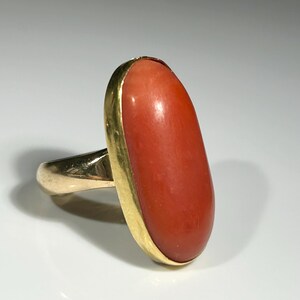 May include: A gold ring featuring a large, oval-shaped coral stone. The coral is a vibrant reddish-orange color, set in a gold bezel. The ring's band is also gold, creating a classic and elegant design.