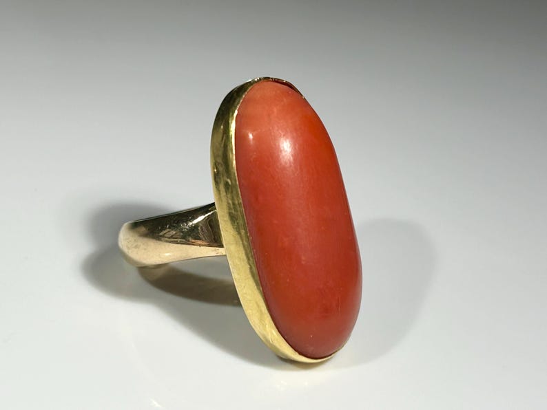 May include: A gold ring featuring a large, oval-shaped coral stone. The coral is a vibrant reddish-orange color, set in a gold bezel. The ring's band is also gold, creating a classic and elegant design.