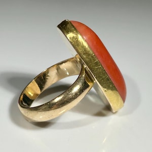 May include: A gold ring with a rectangular, orange-red stone. The stone is set in a gold bezel. The ring band is also gold. The ring is shown on a white surface.
