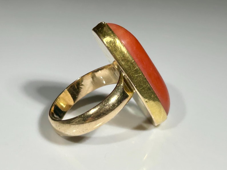 May include: A gold ring with a rectangular, orange-red stone. The stone is set in a gold bezel. The ring band is also gold. The ring is shown on a white surface.
