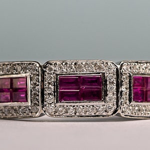 May include: A silver ring with three rectangular ruby stones set in a diamond halo. The rubies are a deep pink color and the diamonds are clear.