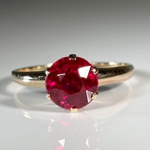May include: A gold ring featuring a large, round, faceted deep red gemstone set in a four-prong mounting. The band is smooth and polished, showcasing a classic design with the vibrant red stone as the central focal point of the jewelry piece.