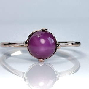 May include: A rose gold ring with a large, round, deep purple gemstone. The gemstone is set in a delicate bezel setting and the band has a simple, elegant design. The ring is displayed on a reflective surface.
