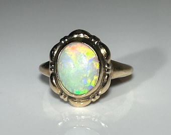 Puede incluir: Un anillo de oro con una piedra preciosa de ópalo ovalada. El ópalo muestra un juego de colores con tonos verdes, amarillos y azules. El anillo tiene un engaste decorativo y festoneado.