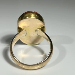 May include: A gold-toned ring with a large, oval-shaped, reddish-orange gemstone. The ring's band is smooth and rounded, and the gemstone is set in a gold bezel.