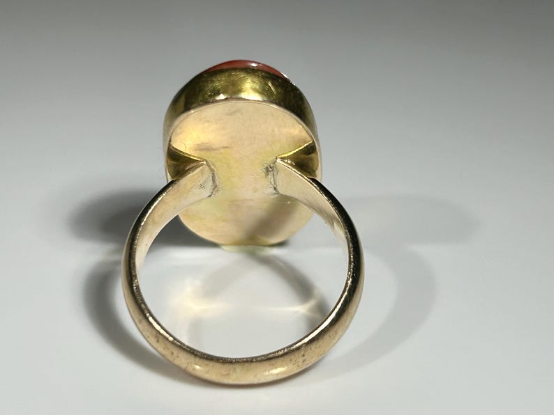 May include: A gold-toned ring with a large, oval-shaped, reddish-orange gemstone. The ring's band is smooth and rounded, and the gemstone is set in a gold bezel.