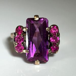 Puede incluir: Un anillo de oro con una gran piedra preciosa púrpura rectangular y facetada en el centro. A ambos lados de la piedra principal, racimos de tres piedras preciosas rosas redondas más pequeñas. El anillo tiene un diseño clásico y elegante.