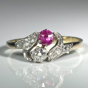 May include: A gold ring featuring a central round-cut pink gemstone and a round-cut clear gemstone, flanked by two curved metal sections set with small clear stones. The band is polished gold, and the setting displays a bypass-style design.