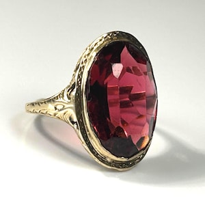 Pode incluir: Um anel dourado com uma grande pedra preciosa vermelha escura, facetada e de forma oval. O anel tem uma banda e uma cravação gravadas e ornamentadas, com um design detalhado em torno da gema. O anel está sobre um fundo branco.