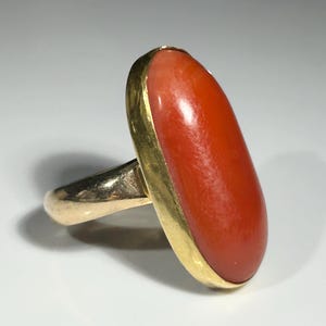 May include: A gold ring featuring a large, oval-shaped coral stone. The coral is a vibrant orange-red color, set in a gold bezel. The ring's band is also gold, creating a classic and elegant design.