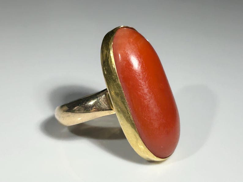 May include: A gold ring featuring a large, oval-shaped coral stone. The coral is a vibrant orange-red color, set in a gold bezel. The ring's band is also gold, creating a classic and elegant design.
