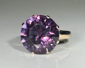 May include: A gold ring featuring a large, faceted, round-cut purple gemstone. The gemstone is held in place by six prongs. The ring's band is a simple, polished gold.