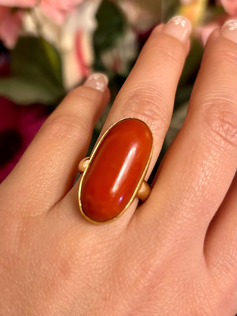 May include: A gold ring featuring a large, oval-shaped, polished red coral stone. The coral is set in a gold bezel, creating a striking contrast. The ring's band is also gold, completing the elegant design. A close-up shot highlights the ring's details.