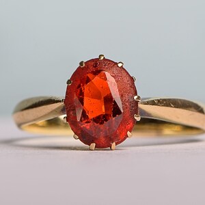 May include: A gold ring with an oval, orange-red gemstone. The gemstone is held in place by small prongs. The ring's band is plain, polished gold. The background is a soft, light blue and white.