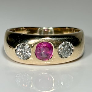 Könnte beinhalten: Ein goldener Ring mit drei Edelsteinen. Der Mittelstein ist ein leuchtend rosa Rubin, flankiert von zwei klaren, funkelnden Diamanten. Der Ring hat einen abgerundeten Schaft und eine polierte Oberfläche.