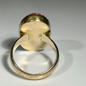 May include: A gold ring with an oval, reddish-orange gemstone. The ring's band and setting are a warm gold color, and the gemstone is smooth and polished. The ring is set against a white background.