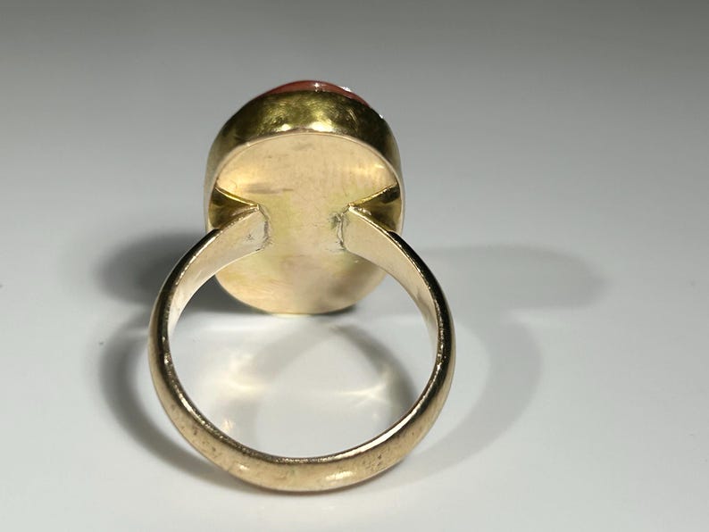 May include: A gold ring with an oval, reddish-orange gemstone. The ring's band and setting are a warm gold color, and the gemstone is smooth and polished. The ring is set against a white background.