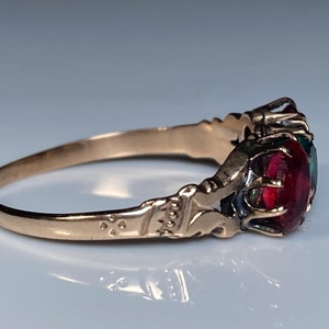 Könnte beinhalten: Ein goldener Ring mit einem markanten, runden, tiefroten Edelstein in einer dekorativen Metallfassung. Das Band weist auf den Seiten komplizierte Gravurdetails auf, die zum zentralen Stein führen und eine polierte metallische Oberfläche zeigen.
