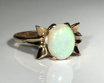 Puede incluir: Un anillo de oro con una piedra preciosa de ópalo ovalada. El ópalo tiene una base blanca lechosa con destellos verdes y amarillos. El engaste del anillo tiene un diseño decorativo y estilizado a ambos lados del ópalo.