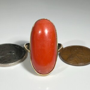 May include: A gold ring featuring a large, oval-shaped, polished coral stone. The coral is a vibrant orange-red color. A coin is on either side of the ring. The ring's band is a simple, classic design.
