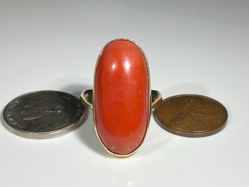 May include: A gold ring featuring a large, oval-shaped, polished coral stone. The coral is a vibrant orange-red color. A coin is on either side of the ring. The ring's band is a simple, classic design.