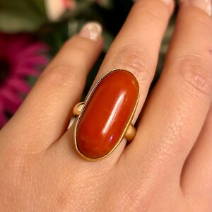 May include: A gold ring featuring a large, oval-shaped, polished red gemstone. The gemstone is set in a simple gold band, creating a classic and elegant design. The ring is shown on a hand, highlighting its size and color.