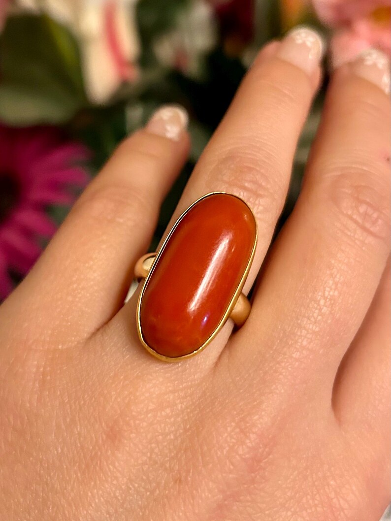 May include: A gold ring featuring a large, oval-shaped, polished red gemstone. The gemstone is set in a simple gold band, creating a classic and elegant design. The ring is shown on a hand, highlighting its size and color.