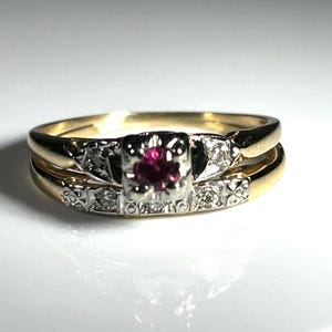 Può includere: Un set di anelli in oro vintage con un rubino centrale e piccoli diamanti di accento. L'anello di fidanzamento ha un'incastonatura quadrata e la fede nuziale è una semplice fascia d'oro. Gli anelli sono esposti su una superficie bianca.