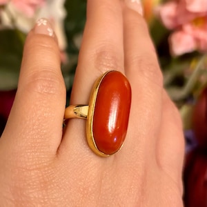 May include: A gold ring featuring a large, oval-shaped, polished red stone. The stone is set in a gold bezel, creating a striking contrast. The ring is a statement piece, suitable for adding a pop of color to any outfit.