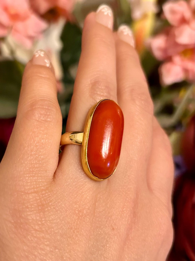 May include: A gold ring featuring a large, oval-shaped, polished red stone. The stone is set in a gold bezel, creating a striking contrast. The ring is a statement piece, suitable for adding a pop of color to any outfit.