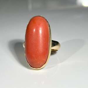 May include: A gold ring featuring a large, oval-shaped coral stone. The coral has a vibrant, reddish-orange hue and is set in a gold bezel. The ring's band is also gold, creating a classic and elegant design.