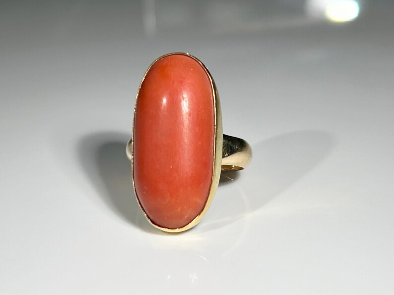 May include: A gold ring featuring a large, oval-shaped coral stone. The coral has a vibrant, reddish-orange hue and is set in a gold bezel. The ring's band is also gold, creating a classic and elegant design.