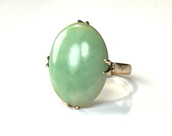 May include: A gold ring featuring a large, oval-shaped, pale green jade stone. The jade is smooth and polished, set in a gold band with claw-like prongs securing the stone. The ring is displayed against a white background.