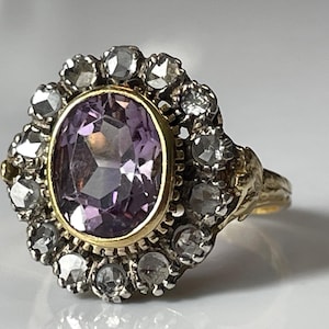 Puede incluir: Un anillo antiguo con una gran amatista ovalada, rodeada de piedras preciosas claras más pequeñas. El anillo tiene una banda y un engaste de oro, mostrando un diseño vintage. Las piedras preciosas brillan, reflejando la luz.