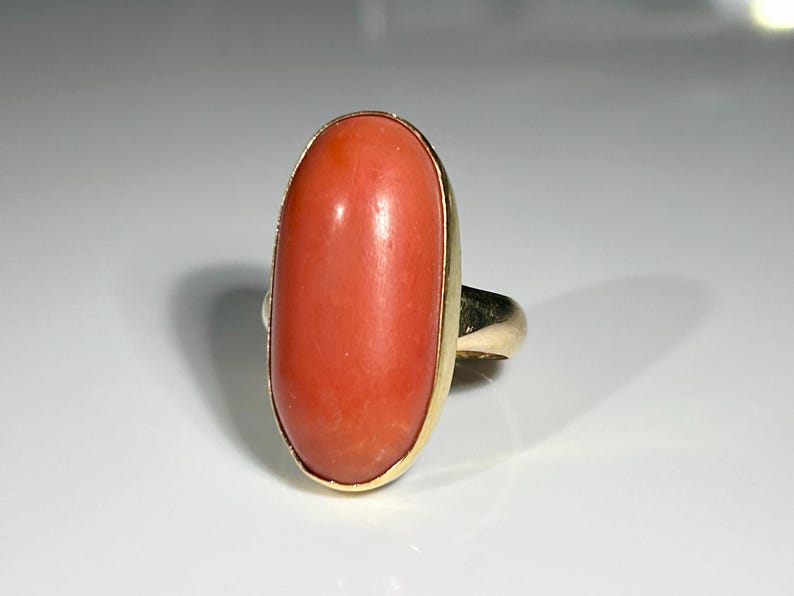 May include: A gold ring featuring a large, oval-shaped coral stone. The coral is a vibrant reddish-orange color, set in a gold band. The ring's design is simple, highlighting the natural beauty of the coral.