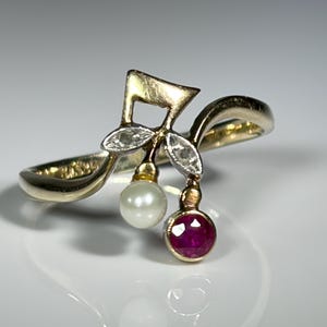 May include: A gold ring featuring a musical note motif accented with two small clear stones, a single white pearl, and a round faceted red ruby. The band has a smooth, polished finish and a curved design that supports the decorative elements.
