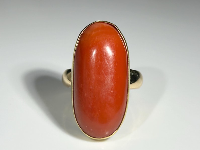 May include: A gold ring featuring a large, oval-shaped, polished coral stone. The coral is a vibrant orange-red color, set in a gold bezel. The ring's band is also gold.