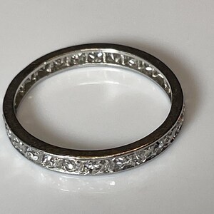 May include: A silver ring featuring a continuous line of small, clear gemstones set around the band. The ring is circular and rests on a white surface, reflecting light.