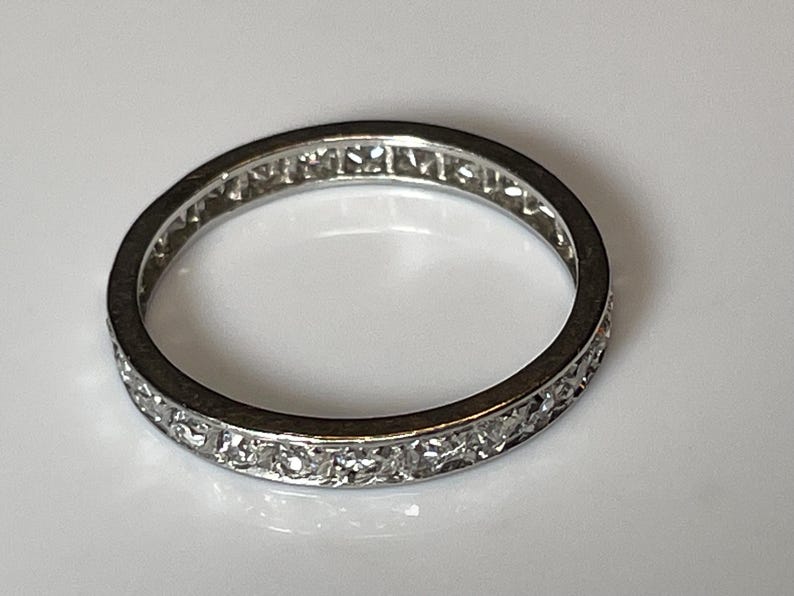 May include: A silver ring featuring a continuous line of small, clear gemstones set around the band. The ring is circular and rests on a white surface, reflecting light.
