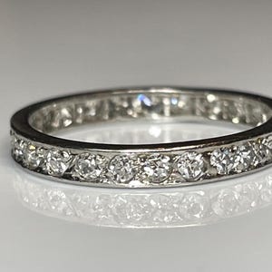 May include: A silver eternity ring featuring a continuous row of round-cut diamonds. The diamonds are set closely together, creating a sparkling band. The ring is displayed on a reflective surface.